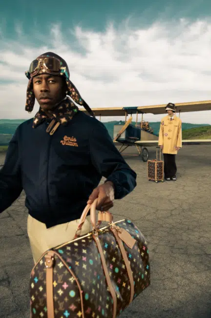 Tyler, the Creator wearing a Louis Vuitton aviator-style outfit with a monogram duffle bag, standing in front of a vintage plane with another model in the background holding matching luggage.