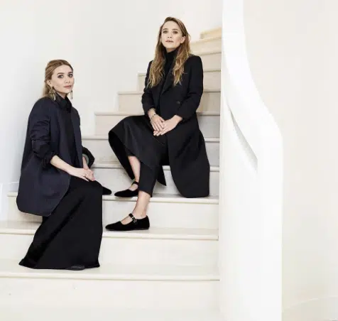 Ashley and Mary-Kate Olsen seated on a curved white staircase, both dressed in elegant long black coats and flowing skirts, embodying minimalist fashion.