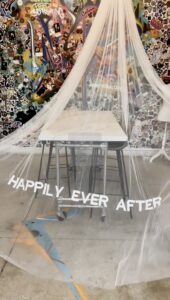 1. Swatch room table covered by a white canopy with “Happily Ever After” text, set against a graffiti-covered wall. A whimsical canopy installation in the swatch room, featuring the phrase “HAPPILY EVER AFTER” draped over a central table.