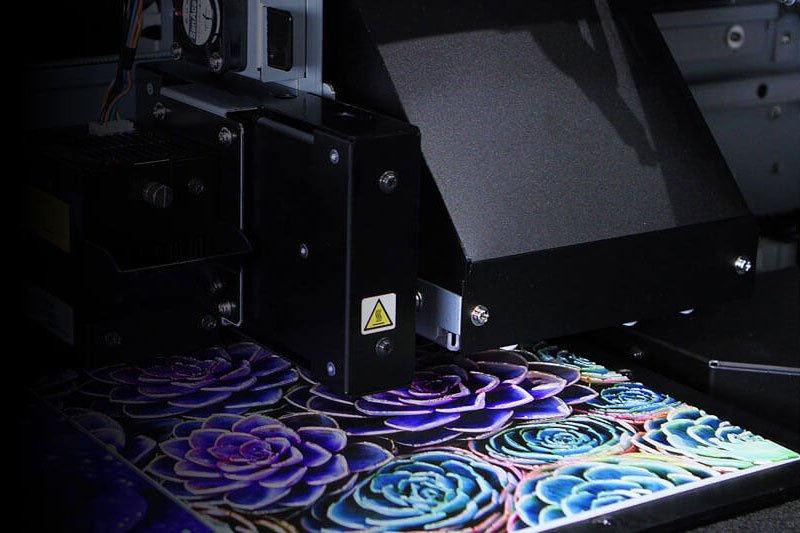 Close-up of a direct-to-garment (DTG) printer printing a vibrant floral design onto fabric, with the printhead moving in reverse.