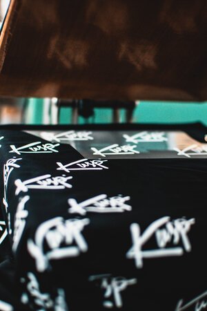 Heat press machine applying a white graphic print with the word “KURP” onto a black fabric.