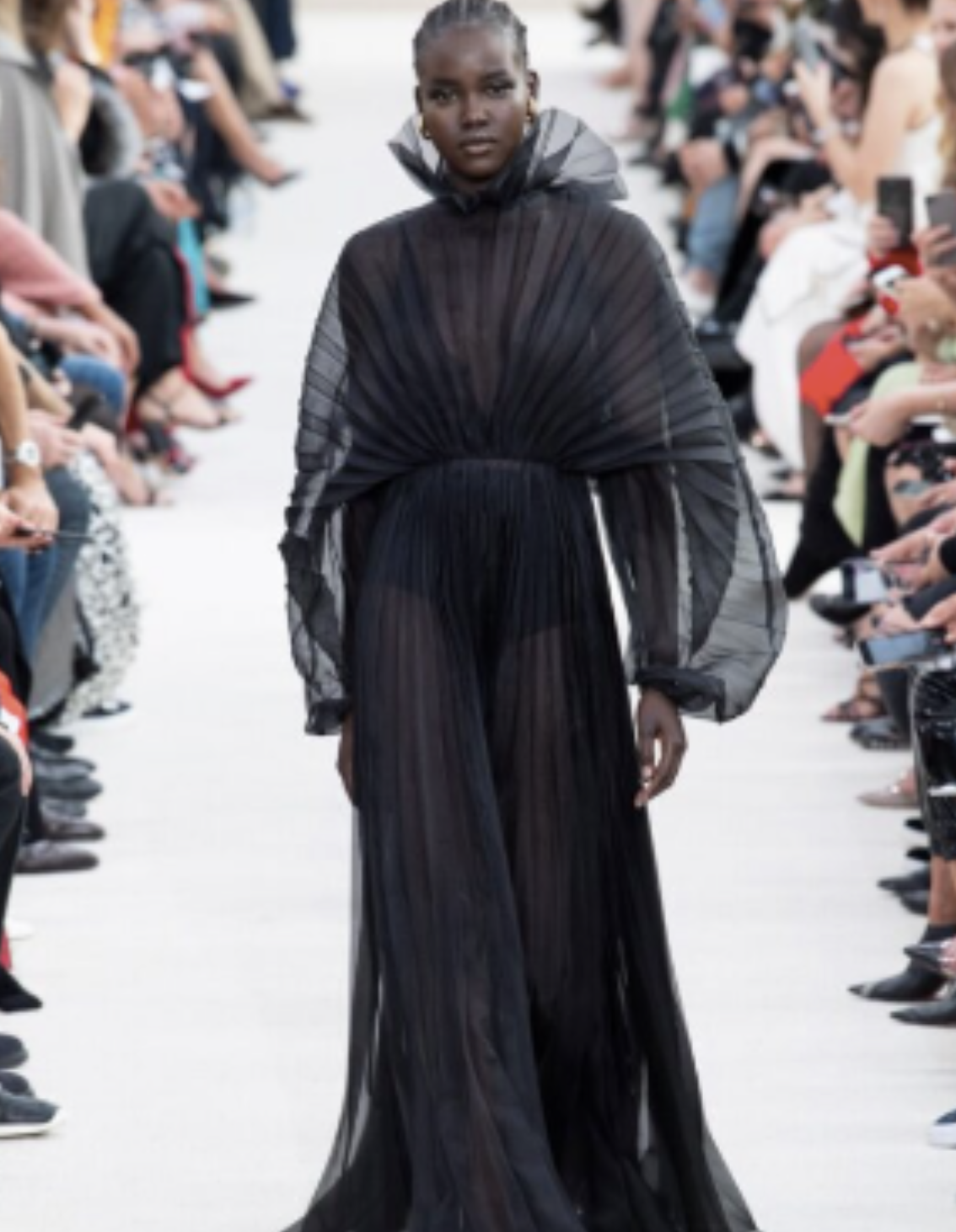 A Black model walks the runway in a sheer black pleated gown with voluminous sleeves and a high ruffled neckline from Valentino’s Spring 2019 collection.