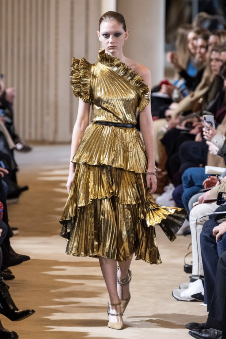 A model walks down the runway in a metallic gold pleated dress with a one-shoulder ruffled neckline and tiered skirt from Altuzarra’s Fall 2019 collection.