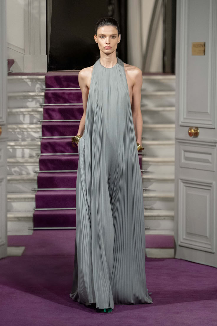A model walks down a runway in a soft grey pleated halter gown by Valentino, with hands tucked into hidden pockets.