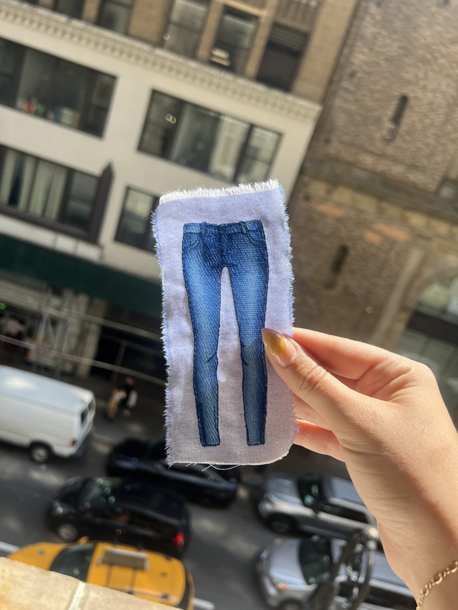A hand holding a fabric swatch embroidered with a hyper-realistic image of blue skinny jeans, with a city street blurred in the background.
