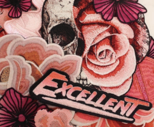 This image features a bold and artistic cluster of embroidered patches. At the center is a standout design: a half skull seamlessly fused with a blooming rose, rendered in rich shades of red, pink, and black — giving it a rebellious yet romantic vibe. Surrounding it are various floral patches in similar warm tones, with detailed stitching and layered textures that add depth and visual interest. The word “EXCELLENT” is prominently embroidered in a retro-inspired font, blending light pink with bold black and coral tones. This patch brings a punchy, graphic element to the mix, giving the whole composition a cool, punk-meets-feminine feel. It’s playful, edgy, and full of personality.