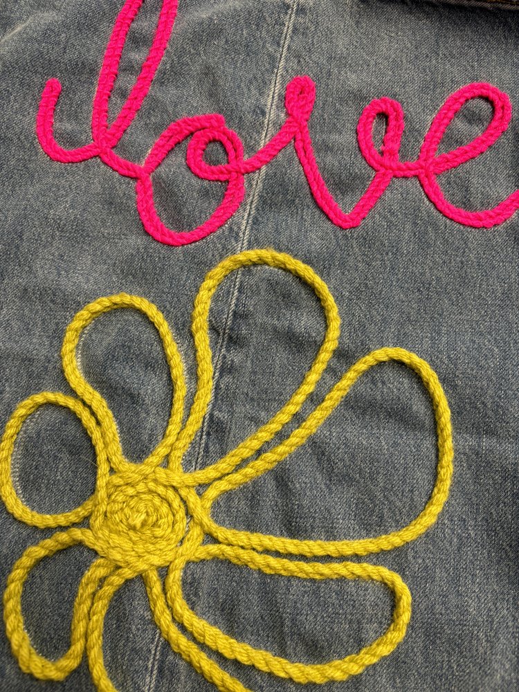 Embroidered denim fabric with the word “love” in pink yarn and a yellow flower motif below, created with chainstitch embroidery.