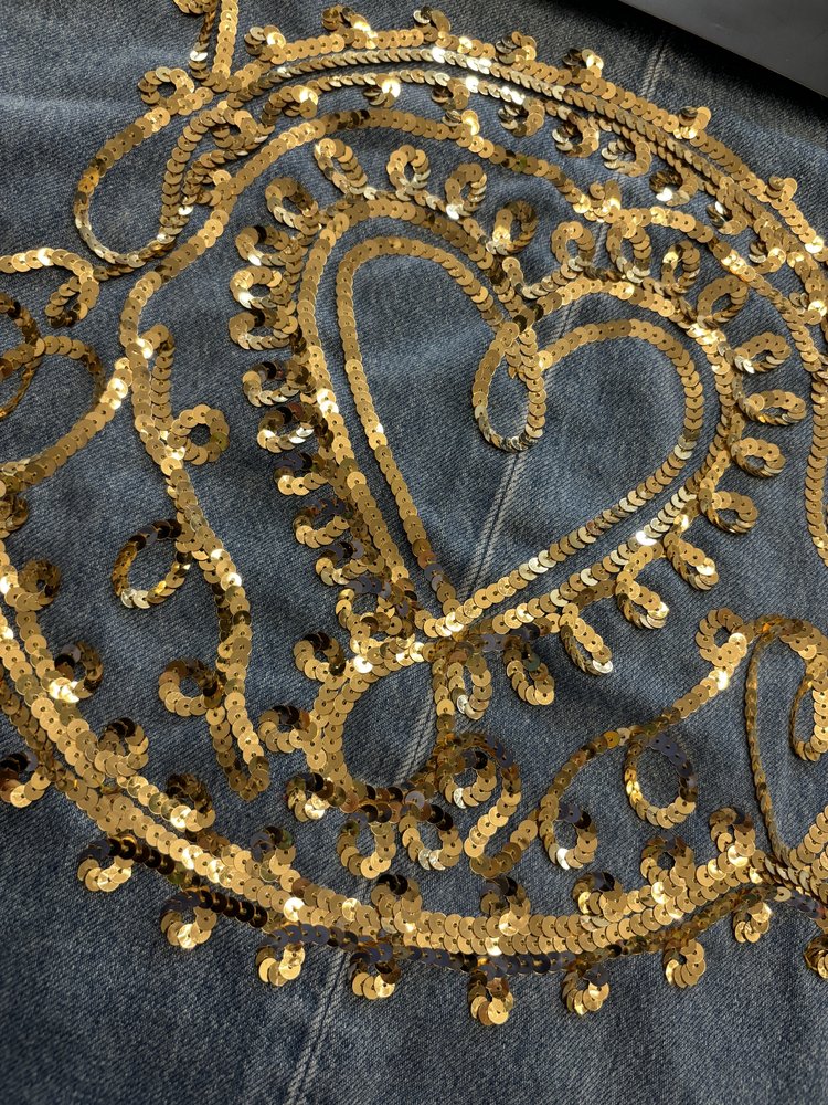 Close-up of a decorative gold sequin embroidery design stitched in a swirling heart motif on blue denim fabric.