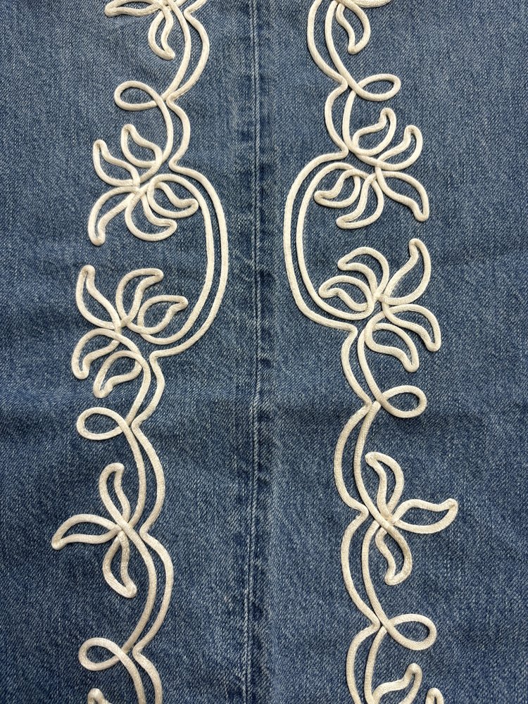 Symmetrical cream cord embroidery stitched onto blue denim, forming mirrored botanical motifs along a center seam.