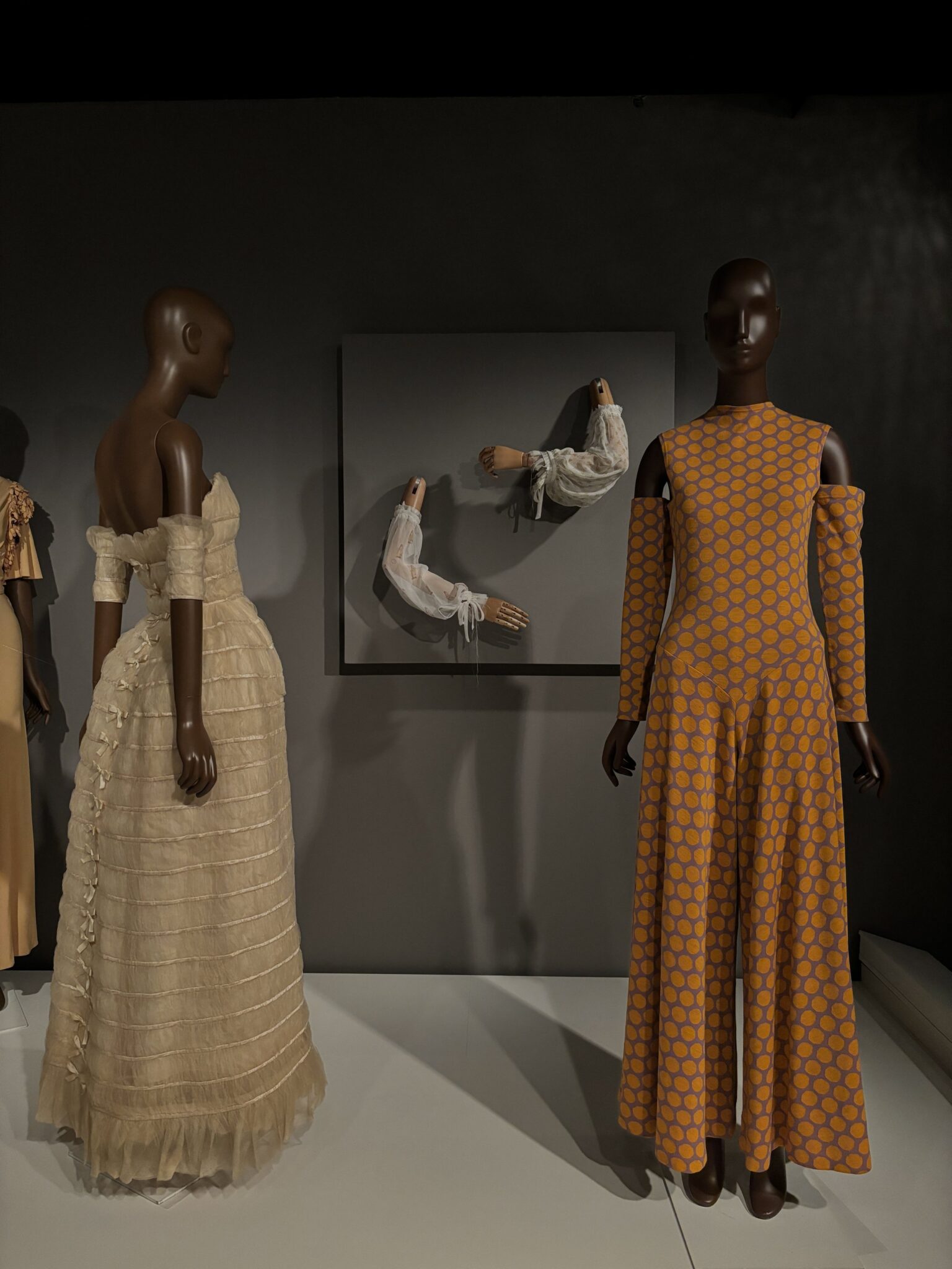Two mannequins display Stephen Burrows designs—a cream pleated off-shoulder gown and an orange patterned long-sleeve jumpsuit—with sculptural white sleeves framed on the wall behind them.