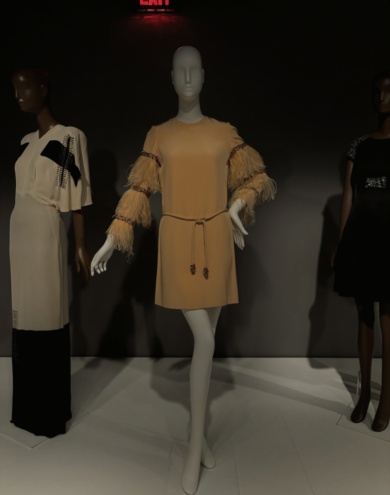 Three mannequins wearing elegant evening dresses, including a beige mini dress with feathered sleeves, a two-tone gown, and a black embellished dress.
