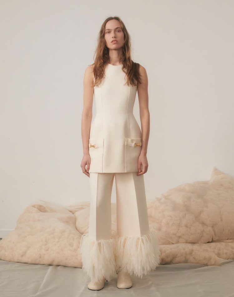 Woman in a sleeveless ivory tunic and matching pants with dramatic feathered hems, standing in a softly lit room with plush furnishings.