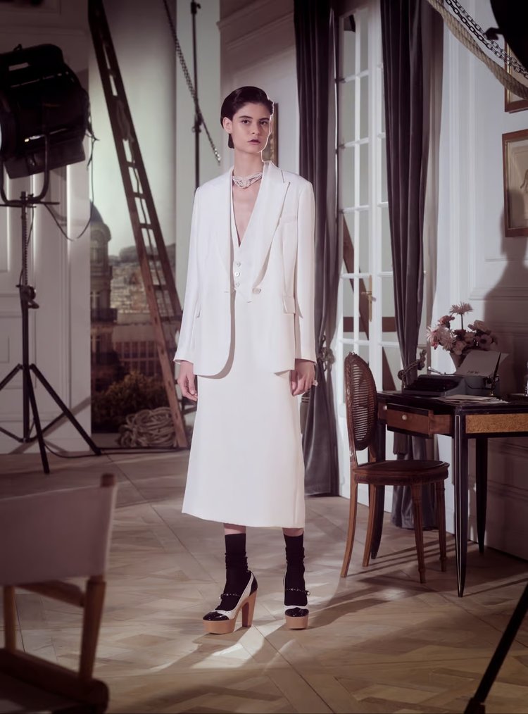 Woman wearing a white tailored blazer and midi skirt with platform heels and black socks, standing in an elegant studio with vintage décor.