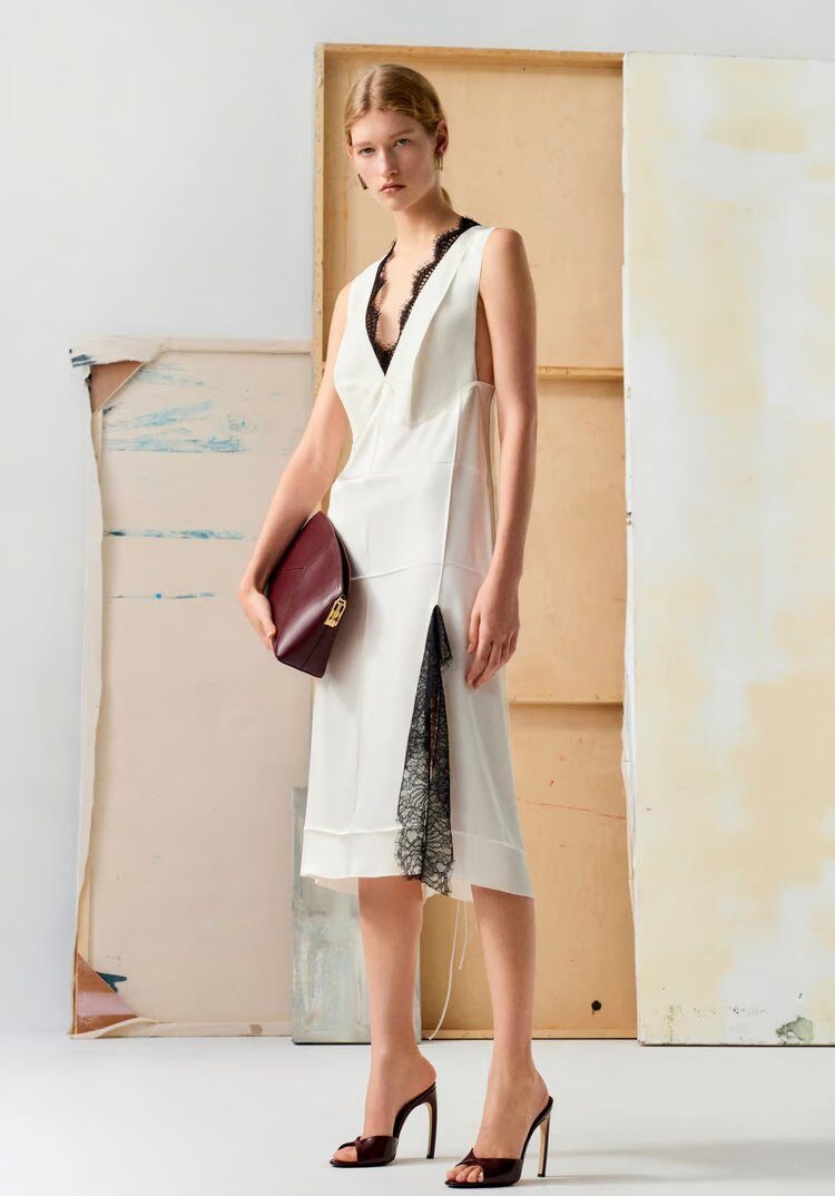 Woman in a sleeveless white V-neck dress with black lace trim, holding a burgundy clutch and wearing black heels, standing in front of a neutral, textured backdrop.