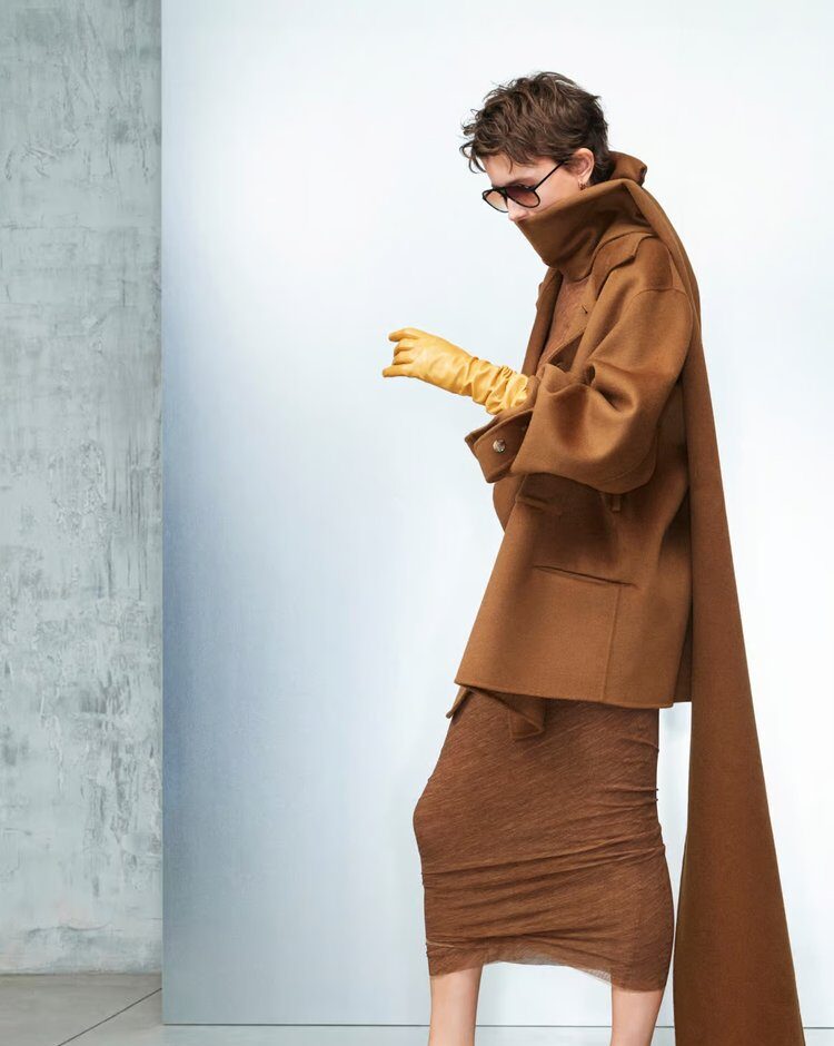 Woman wearing a monochromatic brown outfit with an oversized coat, long scarf, ruched skirt, yellow gloves, and sunglasses.