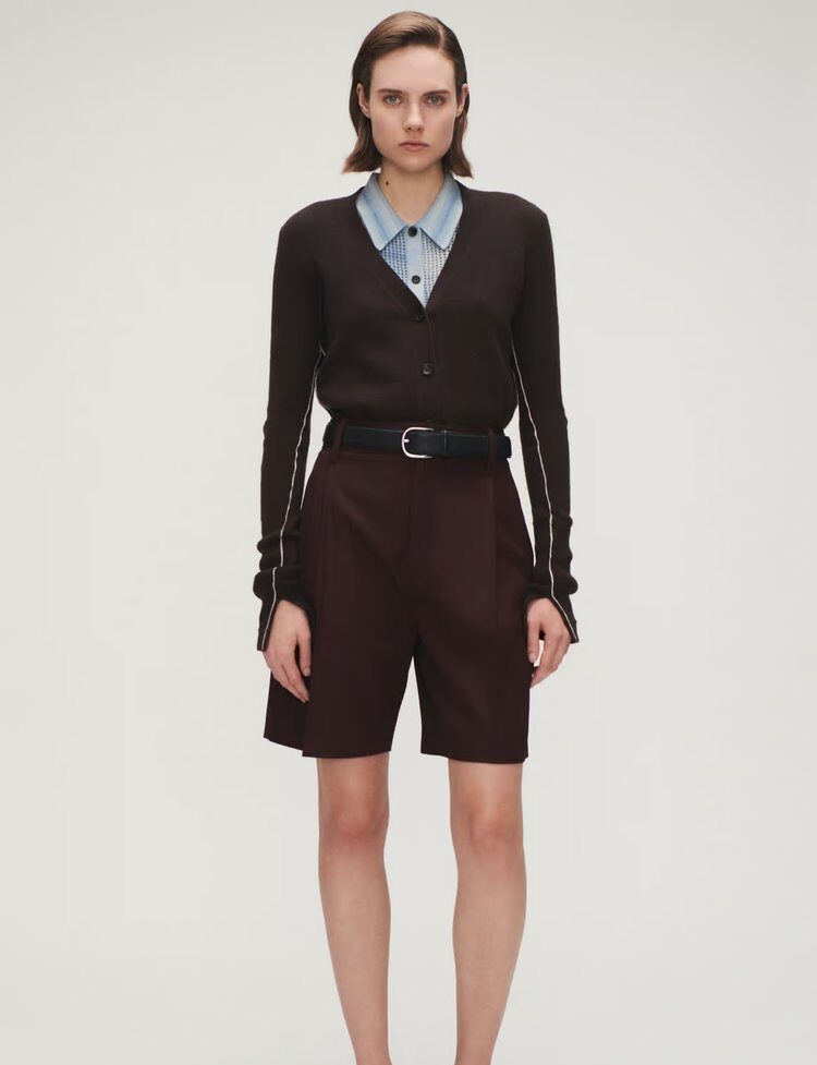 Woman wearing a dark brown cardigan over a blue collared shirt, paired with high-waisted burgundy tailored shorts and a black belt.