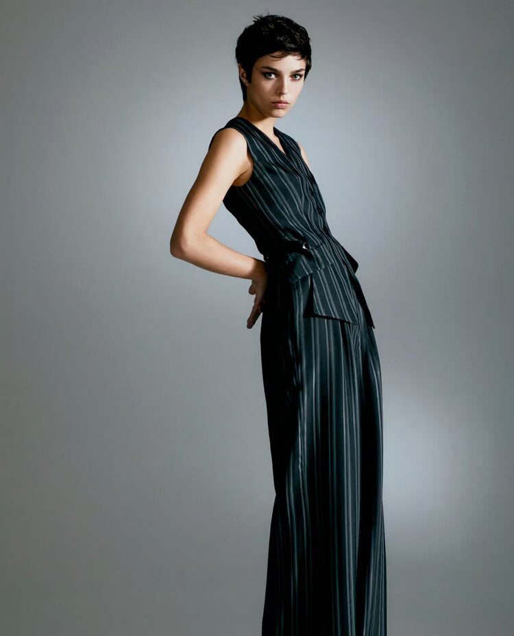 Woman in a sleeveless black pinstripe suit with wide-leg pants, posing against a soft grey studio backdrop.