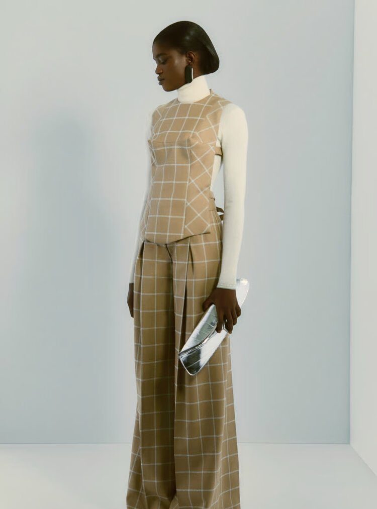 Woman wearing a beige windowpane plaid vest and wide-leg trousers over a white turtleneck, holding a metallic clutch against a pale background.