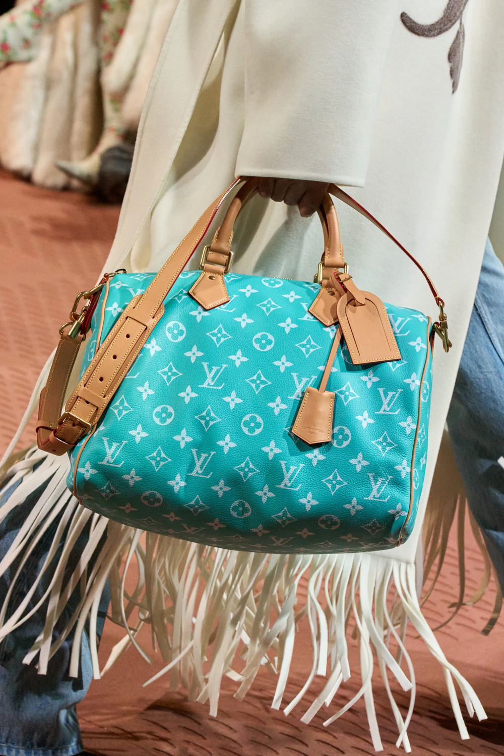 Close-up of a turquoise Louis Vuitton monogram handbag with tan leather straps and gold hardware, worn over a fringed white garment.