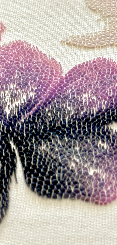 Close-up of beaded embroidery forming a gradient flower in purple, pink, and dark navy shades on a cream textile background.