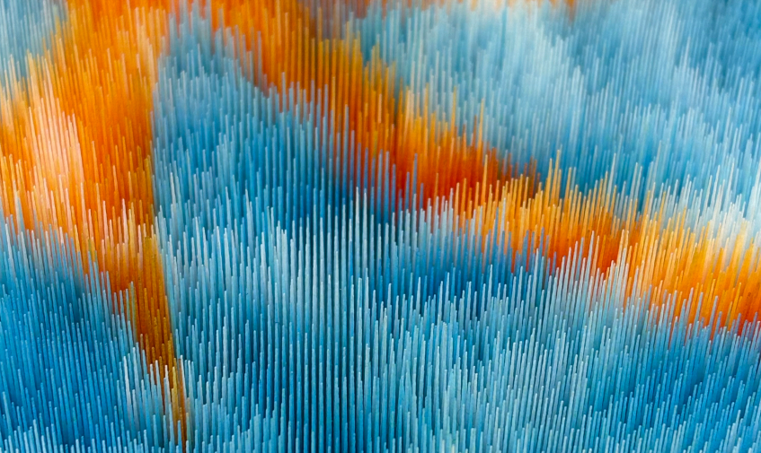 Close-up of a highly textured embroidery piece in vibrant orange, light blue, and dark blue threads, arranged in a directional, brushstroke-like pattern.