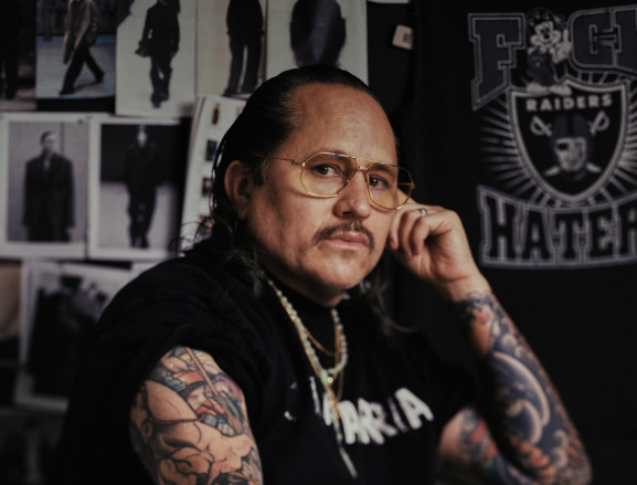 Tattooed designer in glasses seated in front of inspiration wall.