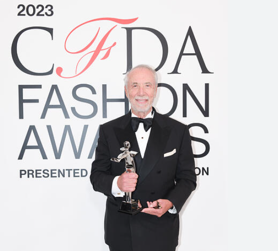 Elderly man in tuxedo holding CFDA trophy on red carpet.