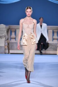 Model walking a runway in a beige dress with satin bow details on the hips.