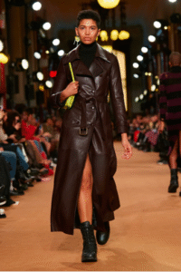 Model walking the runway in a dark brown leather trench coat with a front slit, black turtleneck, combat boots, and a neon green shoulder bag.