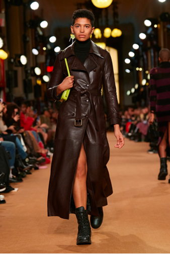Model walking the runway in a dark brown leather trench coat with a front slit, black turtleneck, combat boots, and a neon green shoulder bag.