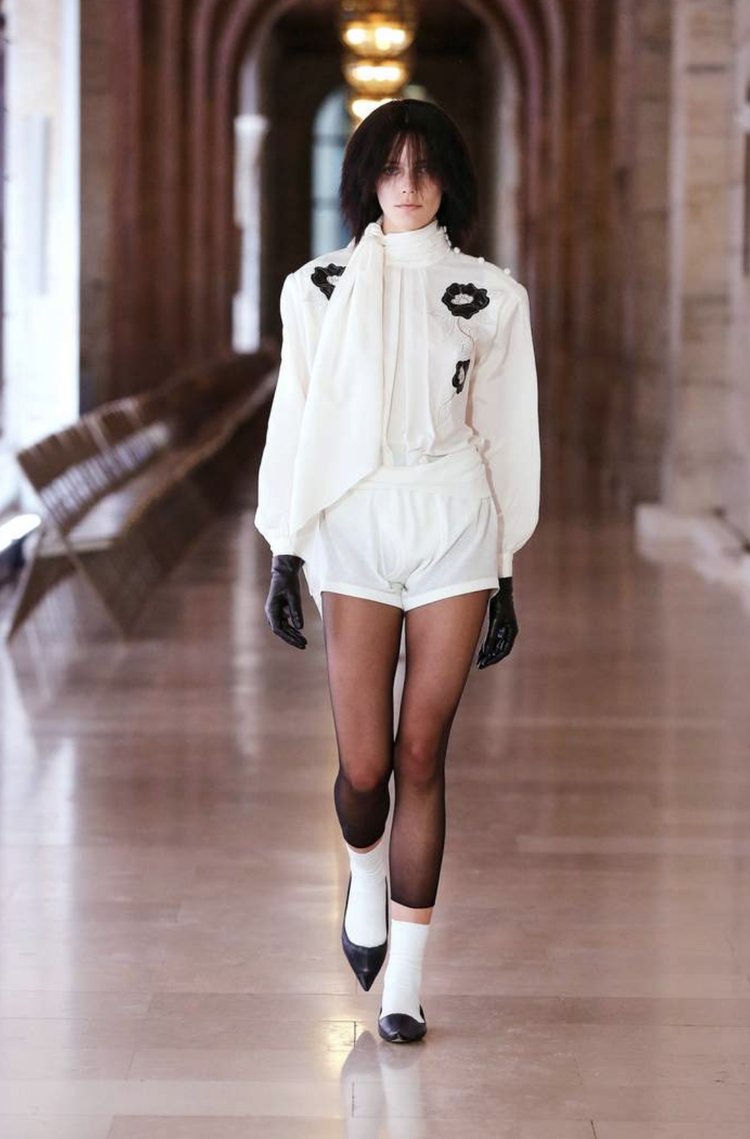 Model walking down a polished hallway wearing an all-white outfit with high-waisted shorts and a structured blouse, styled with black gloves, sheer tights, and pointed heels.