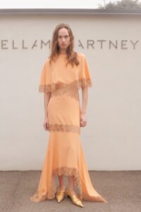 Model stands in flowing peach dress with lace accents and pointed yellow shoes.