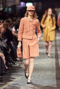 Model wears peach plaid suit with shorts, cap, and matching purse on the runway.