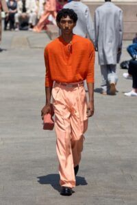 Male model in burnt orange top and satin peach trousers walks outdoors.