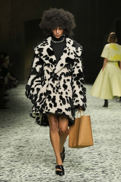 Model walking in black and white cow-print fuzzy coat, black turtleneck, and neutral tote.
