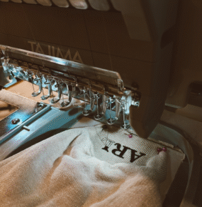 Close-up of an embroidery machine stitching the initials “AR” on beige fabric.