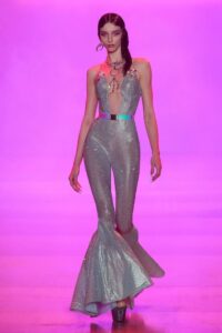 Model wearing a metallic silver jumpsuit with flared hems and a halter neckline walking a runway with a vivid pink-purple background at NYFW Spring/Summer 2023.