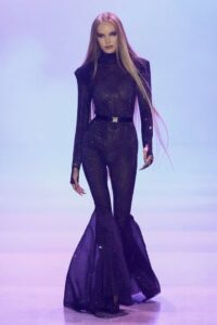 Model in a sheer dark purple jumpsuit with flared legs, strong shoulders, and a belted waist walking a softly lit purple runway at NYFW Spring/Summer 2023.