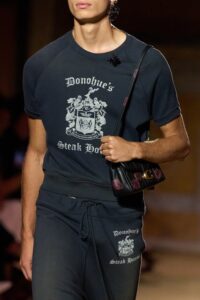 Model wearing a matching navy blue graphic t-shirt and jogger set with “Donohue’s Steak House” printed on both pieces, accessorized with a small crossbody bag.