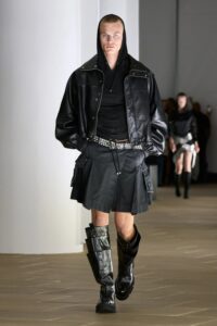 Model walking in a black mini pleated skirt and oversized black leather jacket, styled with a belted waist and tall boots.