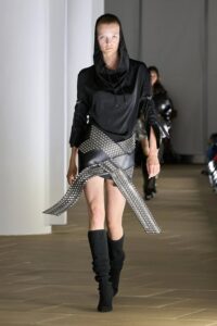 Model in a black asymmetrical top and metallic mini skirt with crossed belts or straps, styled with black knee-high boots.