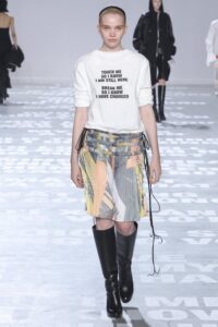 Model wearing a white t-shirt with black text and a semi-sheer printed skirt, paired with black knee-high boots.