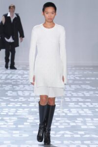 Model in a fitted white long-sleeve knit dress with drawstring details at the hem, styled with black knee-high boots on the runway.