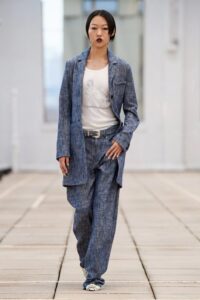 Model walking in a slouchy blue denim-style pantsuit with a white printed tank top and a silver-buckled belt.