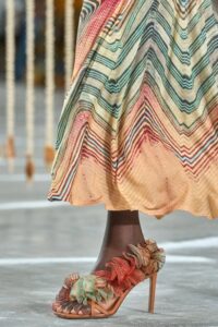Close-up of a model's foot in a strappy coral-toned heeled sandal with multi-colored floral detailing, paired with a pastel chevron-patterned skirt.