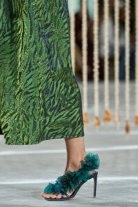 Close-up of a model wearing green and navy open-toe heeled sandals with textured pompom detailing, paired with a green animal-print midi skirt.