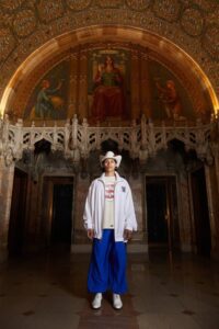Model in a white track jacket, graphic tee, oversized royal blue pants, and white sneakers, accessorized with a large black-and-white hat, standing under a gilded vaulted ceiling.