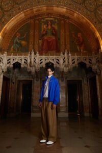 Model wearing a bright blue zip-up jacket, white shirt, and wide-leg camel trousers, standing in a grand hall with arched ceilings and ornate murals.