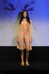 Model wearing a sheer yellow lace robe over orange lingerie on a dark runway.