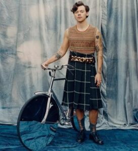 Harry Styles posing in a pleated skirt and knitted vest next to a vintage bicycle.